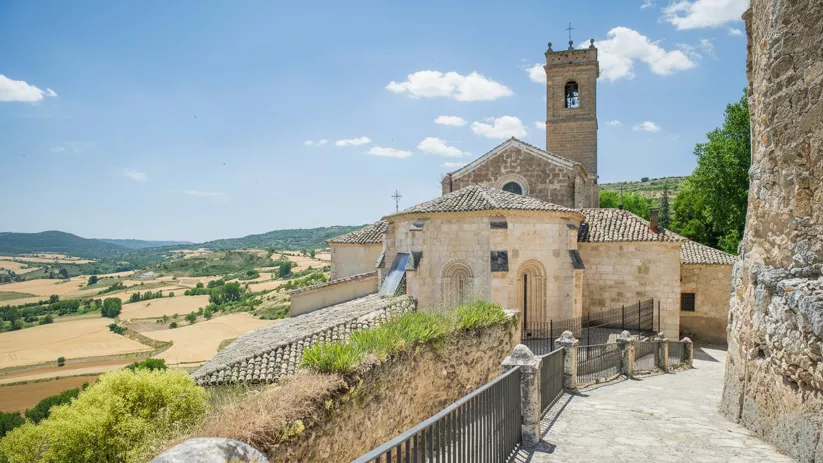 Iglesia de piedra con torre campanario y paisaje de campos ondulados al fondo.