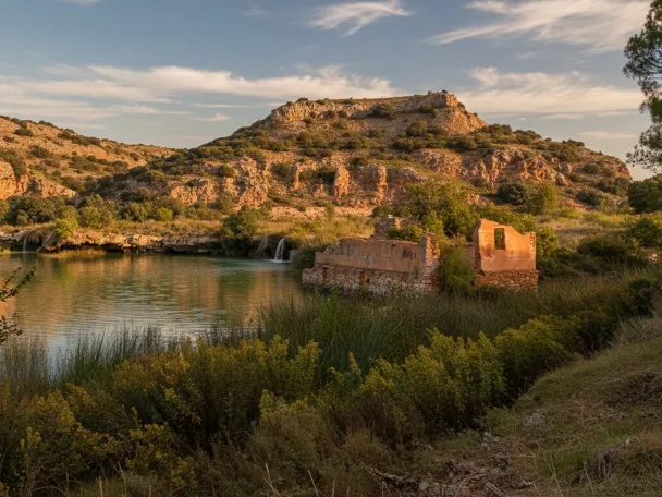Lago tranquilo al atardecer con colinas rocosas y ruinas junto a la orilla.