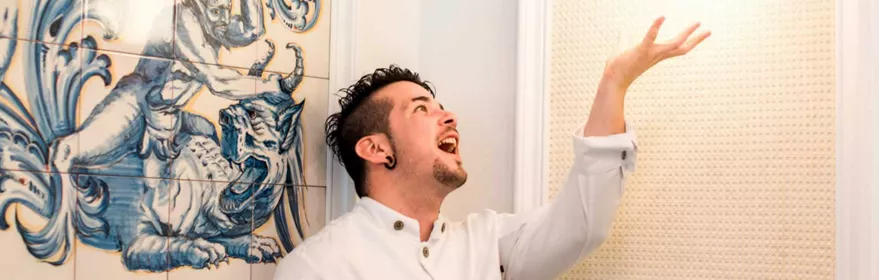 Cocinero sonriendo y alzando la mano junto a mural de azulejos