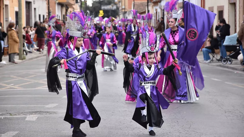Comparsa infantil con trajes morados y estandarte durante un desfile festivo