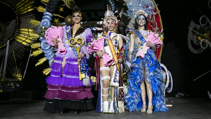 Tres personas con trajes coloridos y abanicos posan sobre un escenario durante un desfile.