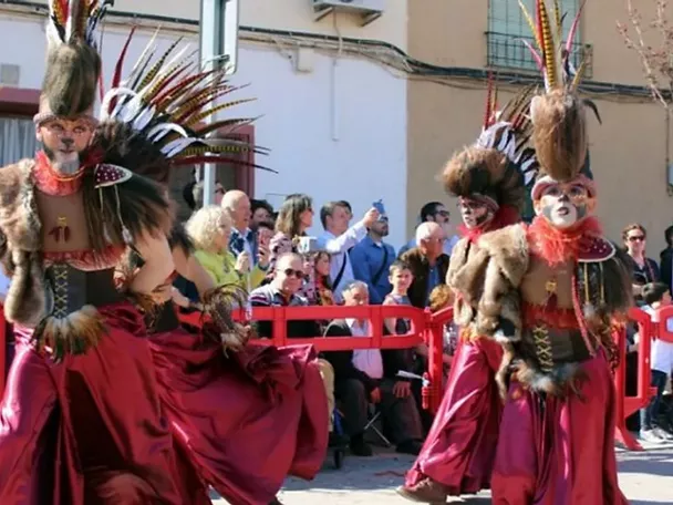 Dos personas con disfraces tribales, plumas altas y faldas rojas desfilando ante el público.