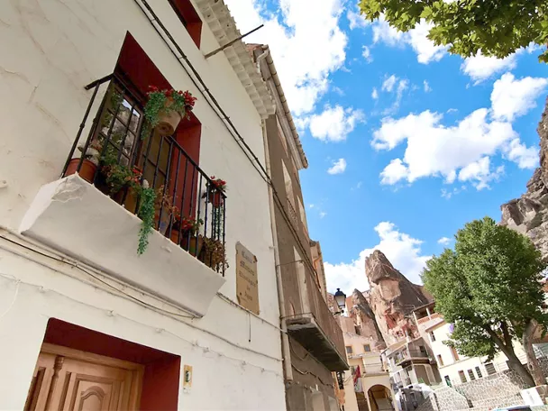 Calle estrecha con fachadas blancas, balcones con plantas y cielo azul entre rocas.