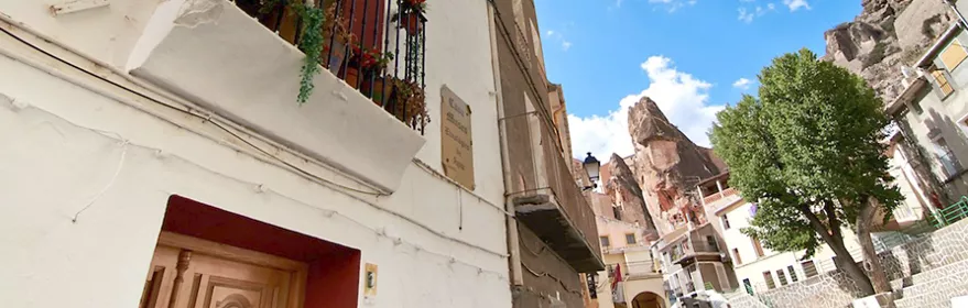 Calle estrecha con fachadas blancas, balcones con plantas y cielo azul entre rocas.