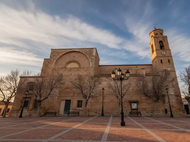 Iglesia de piedra frente a una plaza amplia, con campanario y farolas bajo cielo nublado.