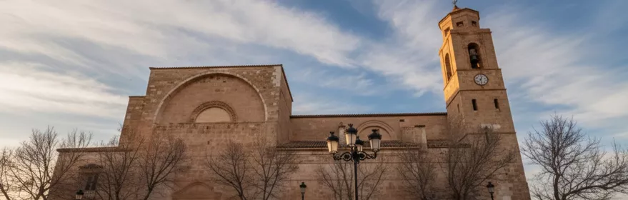 Iglesia de piedra frente a una plaza amplia, con campanario y farolas bajo cielo nublado.