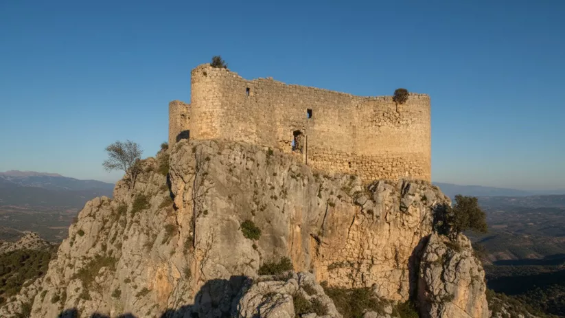 Murallas de piedra sobre una cima rocosa con paisaje montañoso al fondo.
