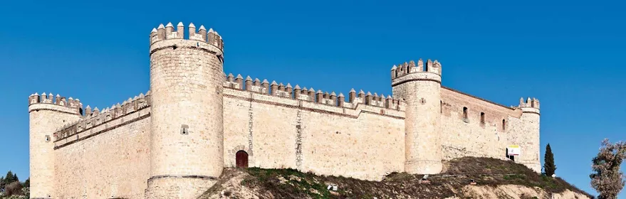 Castillo de piedra con torres circulares y almenas sobre una colina bajo cielo azul.