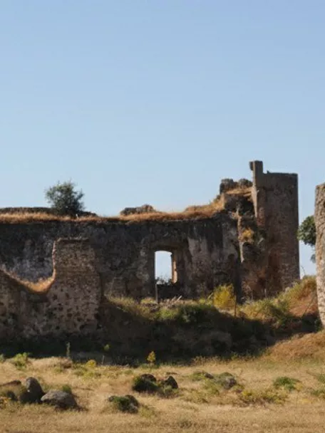 Vista lateral de fortaleza en ruinas sobre terreno seco con torres cilíndricas y cielo despejado.