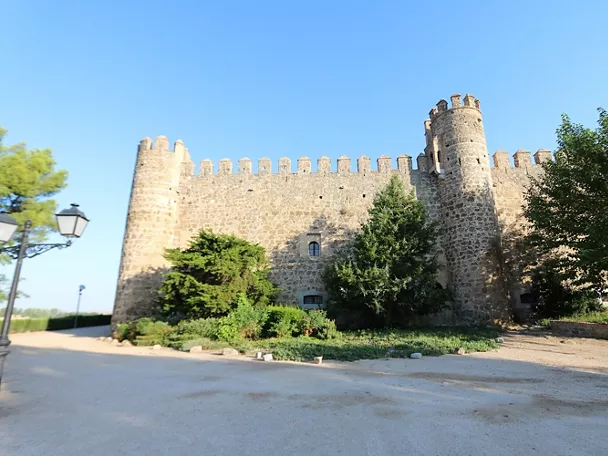 Fachada de piedra con torres y almenas junto a zona ajardinada.