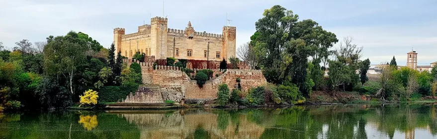 Edificio fortificado de estilo medieval junto al río con murallas y torres cuadradas.