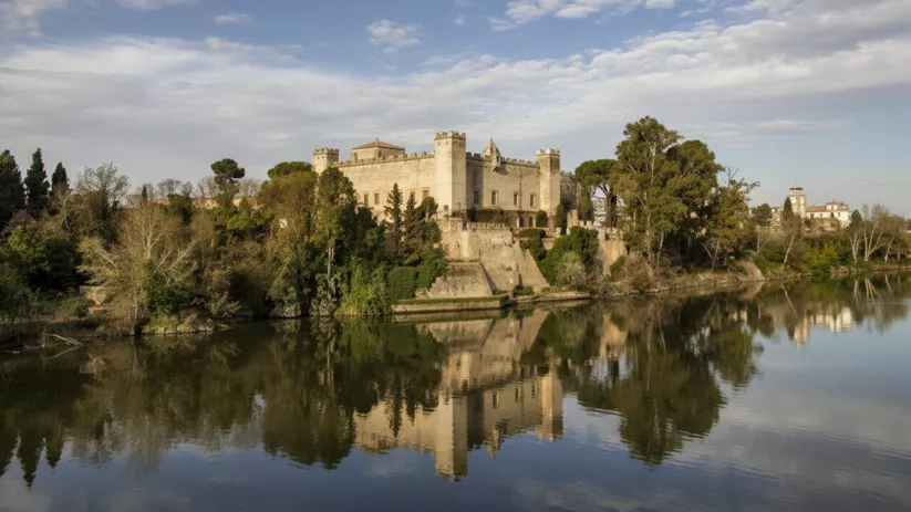 Castillo amurallado sobre la ribera con vegetación densa y reflejo en el río.