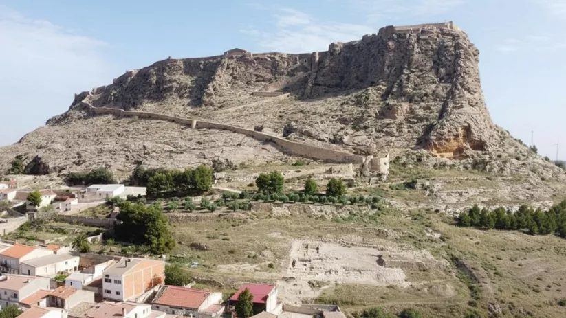 Fortaleza sobre un cerro rocoso, con muralla serpenteante y casas en primer plano.