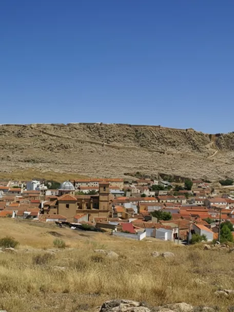 Pueblo con tejados rojizos frente a una gran meseta rocosa coronada por murallas.