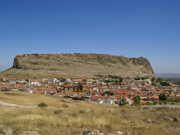 Pueblo con tejados rojizos frente a una gran meseta rocosa coronada por murallas.