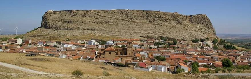 Pueblo con tejados rojizos frente a una gran meseta rocosa coronada por murallas.