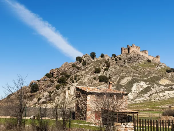 Castillo en lo alto de una colina rocosa con casa rural en primer plano.