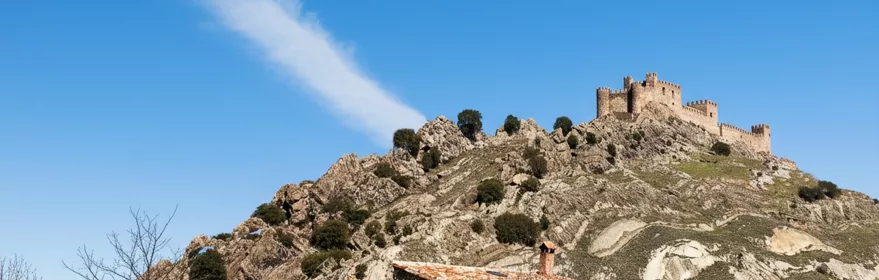 Castillo en lo alto de una colina rocosa con casa rural en primer plano.