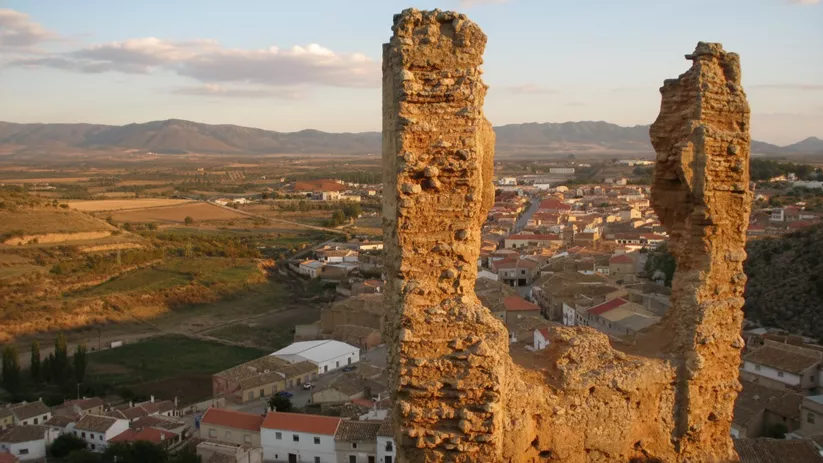 Fragmento de fortificación de piedra sobre el casco urbano, con valle y sierras al atardecer.