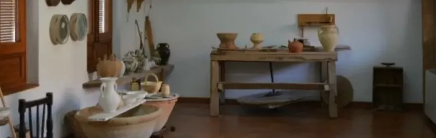 Sala con mesa de trabajo y piezas de alfarería expuestas en estantes.