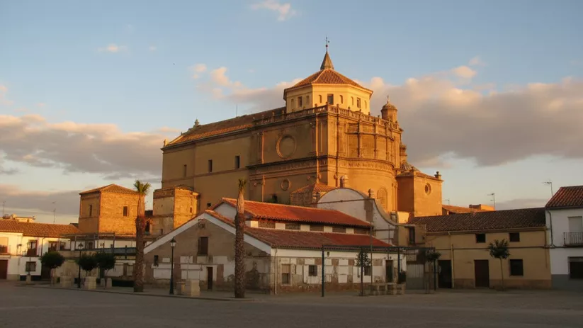 Iglesia de gran volumen vista desde una plaza, iluminada por luz cálida y cielo con nubes.