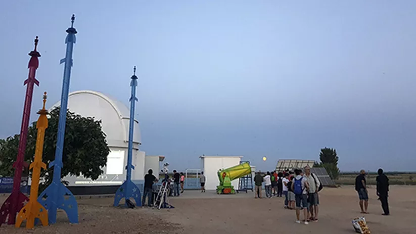 Grupo de personas observando el cielo al atardecer junto a telescopios y estructuras con forma de cohete.