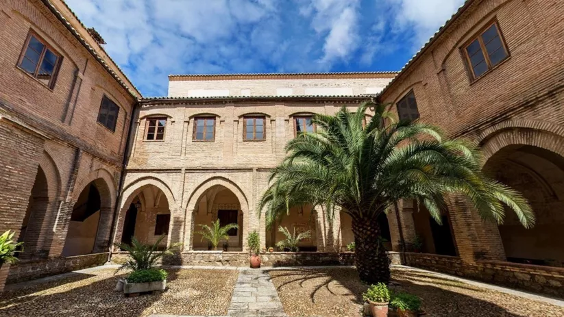 Claustro rectangular con arcos de ladrillo, patio empedrado y palmera central.