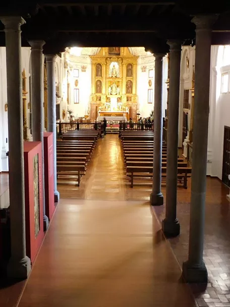 Interior de iglesia con columnas, bancos de madera y altar dorado al fondo.