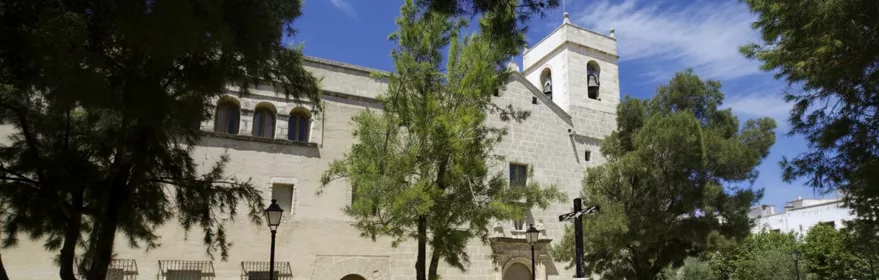 Fachada de iglesia con torre campanario y plaza arbolada.