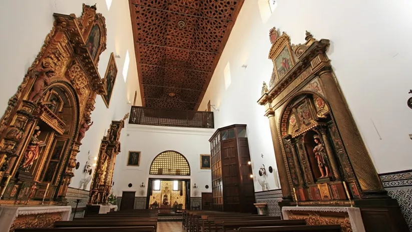 Interior de iglesia con retablos dorados, techo artesonado y bancos de madera.