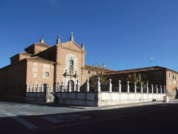 Fachada de iglesia de ladrillo con cruz superior y verja de piedra junto a la calle.