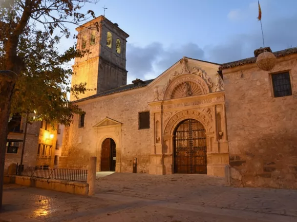 Iglesia parroquial iluminada al atardecer, fachada de piedra y torre campanario en una plaza histórica.