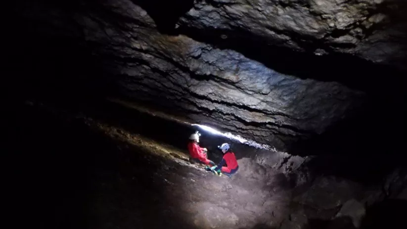 Dos personas con casco y frontal descansan en una galería estrecha dentro de una cueva.