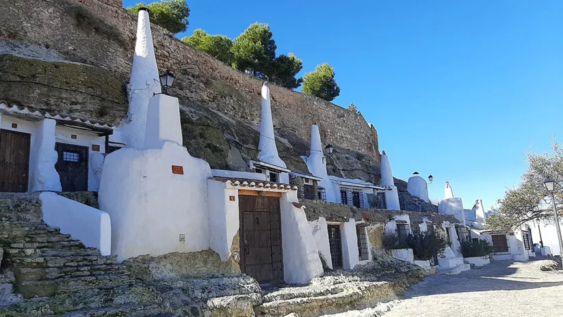 Vista lateral de casas cueva blancas alineadas bajo una muralla de piedra.
