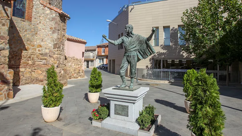 Escultura de figura humana con brazos extendidos en una plaza.