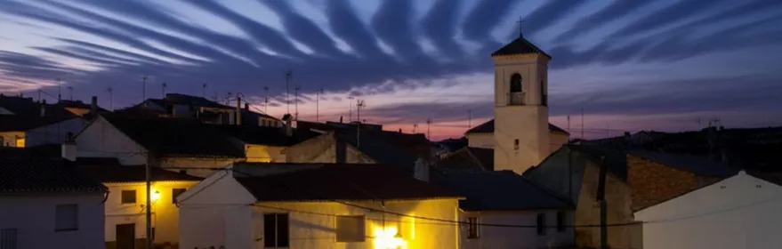 Vista nocturna del pueblo, con casas encaladas iluminadas y una torre sobresaliendo bajo un cielo azul intenso.