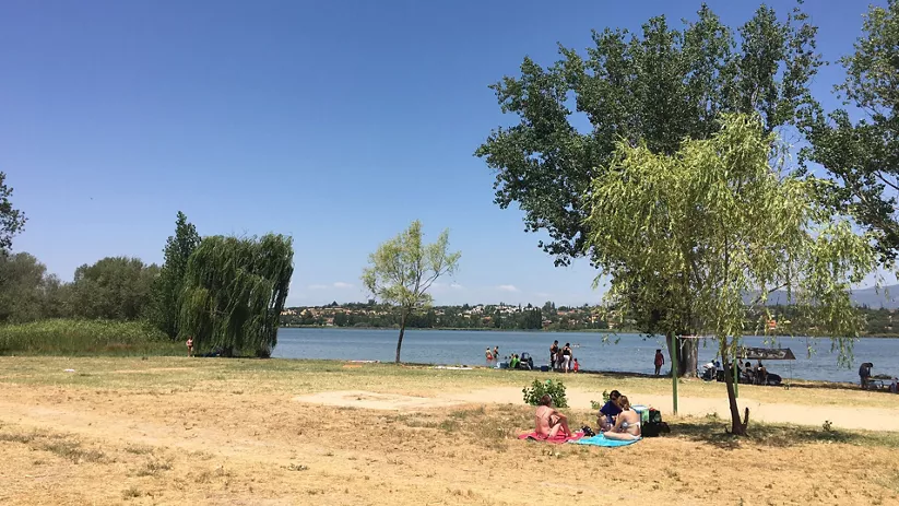 Zona de baño natural junto al embalse con árboles y personas descansando