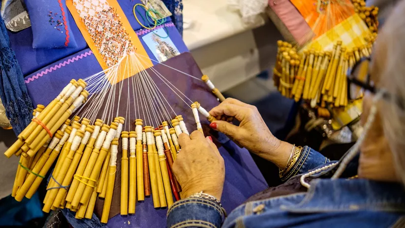 Manos realizando encaje artesanal con bolillos sobre un cojín