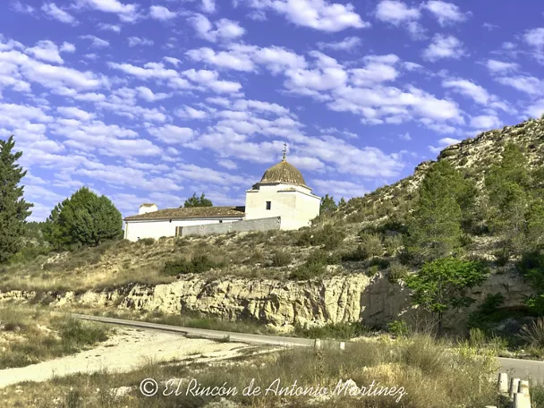 Ermita blanca sobre una ladera rocosa rodeada de vegetación y cielo azul con nubes.