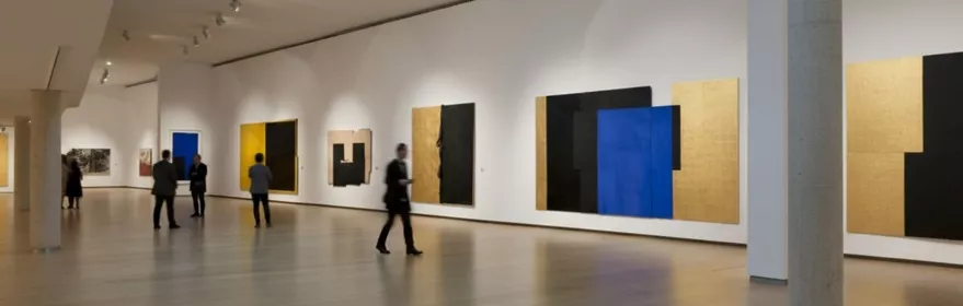 Amplia sala con lienzos minimalistas y visitantes observando.
