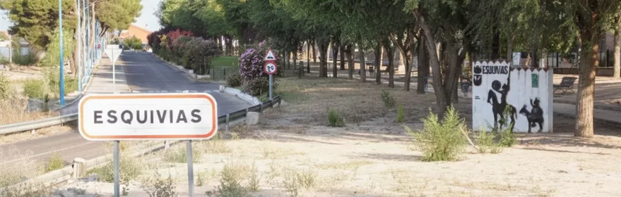 Acceso urbano con señalización y arbolado junto a la carretera.