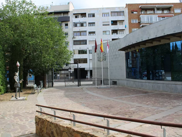 Plaza urbana con edificio cultural moderno, zona ajardinada y banderas