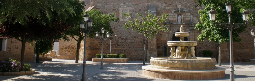 Plaza principal de Fernán Caballero (Ciudad Real) con fuente ornamental y fachada de la iglesia parroquial al fondo.