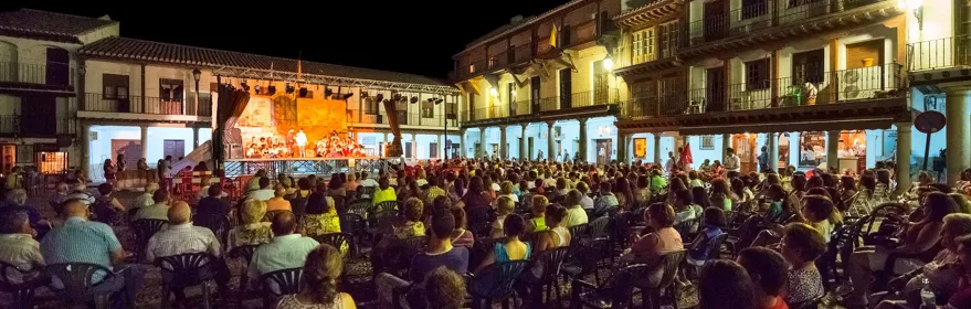 Plaza mayor de noche con escenario iluminado y numeroso público sentado frente a la representación.