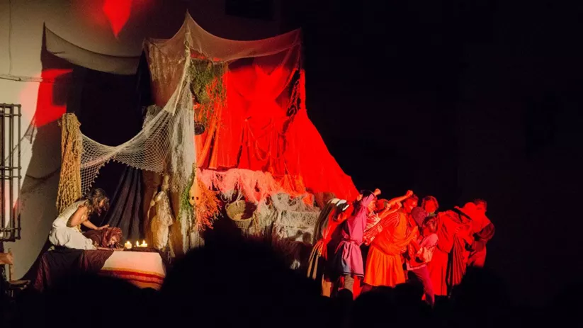 Representación teatral nocturna con grupo de actores bajo iluminación roja y escenografía con telas y redes.