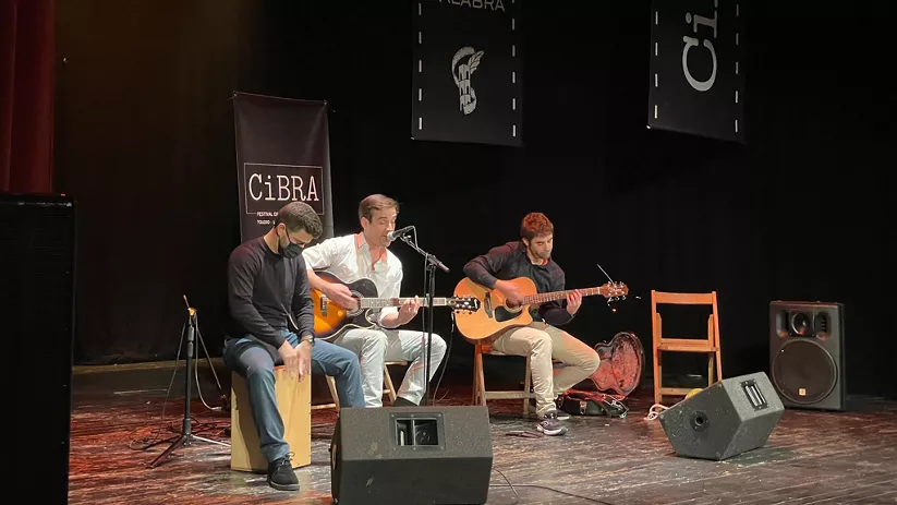 Tres músicos actúan en directo con guitarras y percusión en un escenario