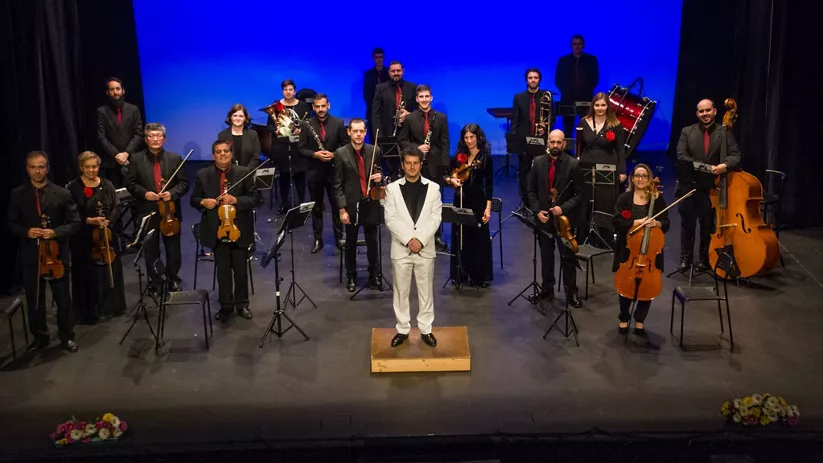 Orquesta sobre escenario con director al frente y músicos con instrumentos.