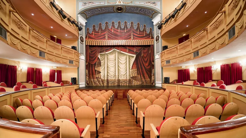 Interior de teatro clásico con escenario frontal, butacas de madera y palcos laterales decorados.