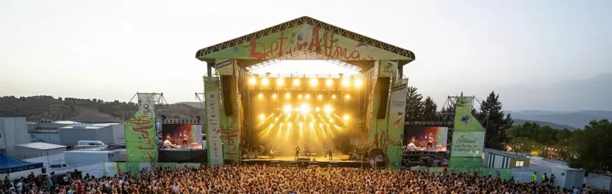 Multitud frente a un gran escenario iluminado durante un concierto al atardecer.