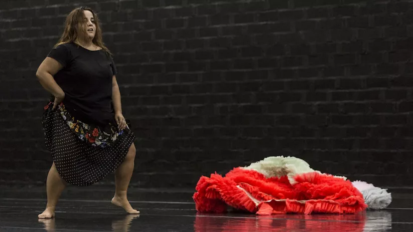 Mujer descalza en un escenario oscuro mirando al frente, con un montón de tela roja a un lado.